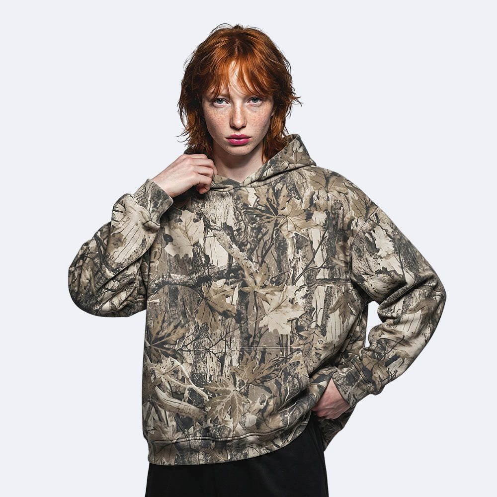 Birch. A model with red hair wearing a camouflage hoodie with a realistic leafy branch pattern in shades of brown, beige, and gray.