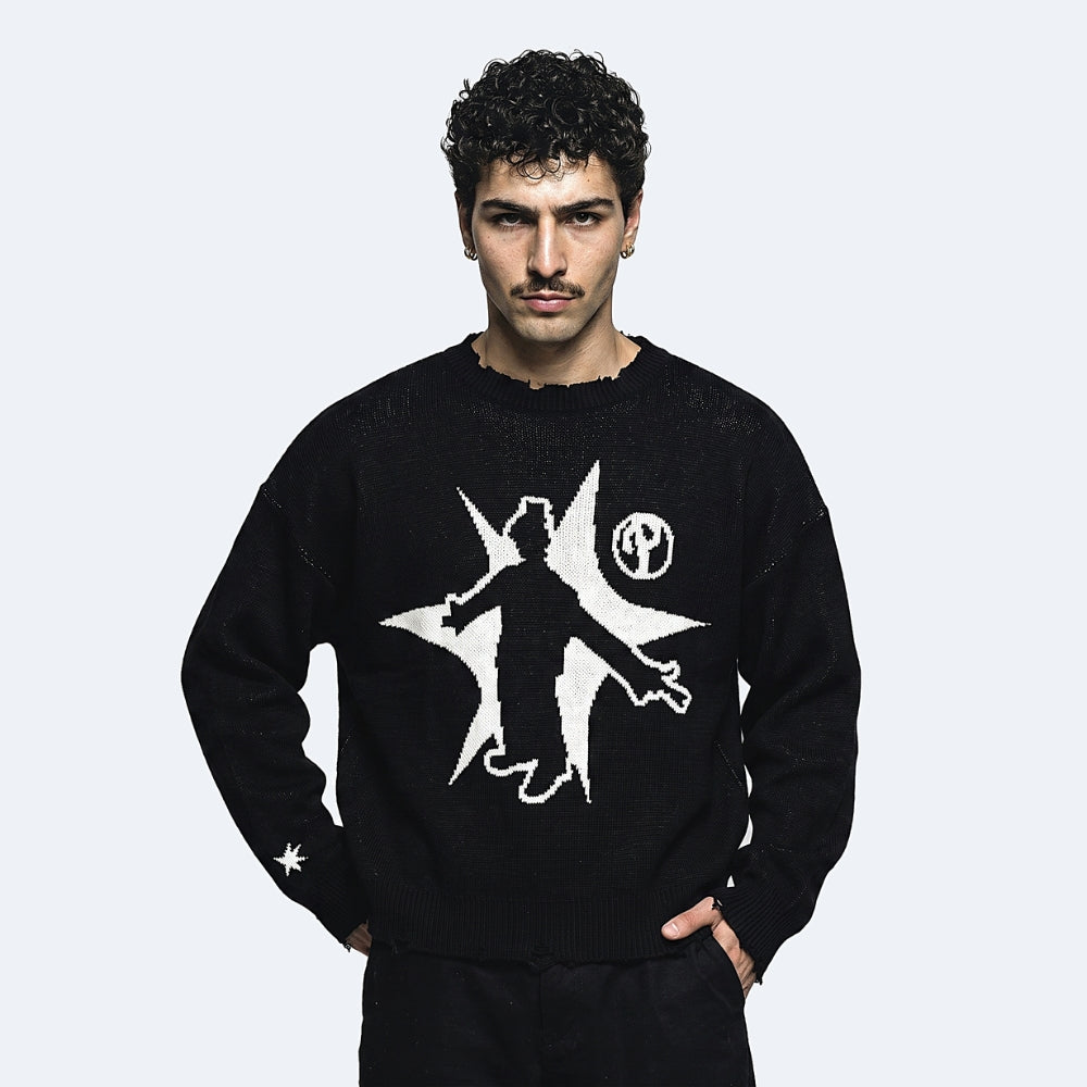 A man wearing a black knit crewneck sweater with a large white graphic of a silhouette inside a starburst and a circular emblem on the chest, posed against a light gray background.