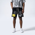 A model is shown from the waist down wearing black washed athletic shorts with white drawstrings, a yellow 'TERRITORY OF THE LAP NINE' patch on the left leg, and white sneakers, paired with a gray graphic t-shirt against a light gray studio background.