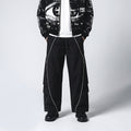 A model wearing black wide-leg cargo pants with white piping, a white t-shirt, and a black graphic zip-up jacket against a light gray studio background.