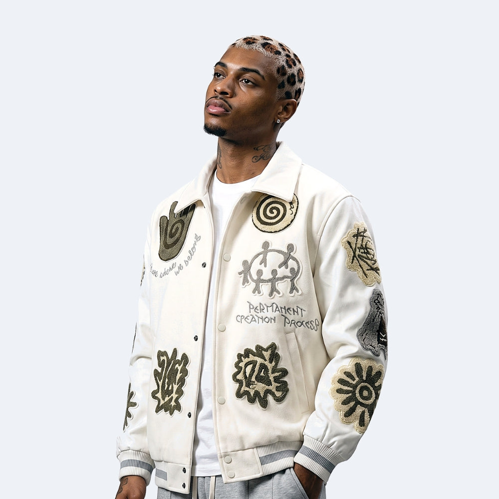 White. A man wearing a cream varsity-style bomber jacket with multiple embroidered patches and designs.