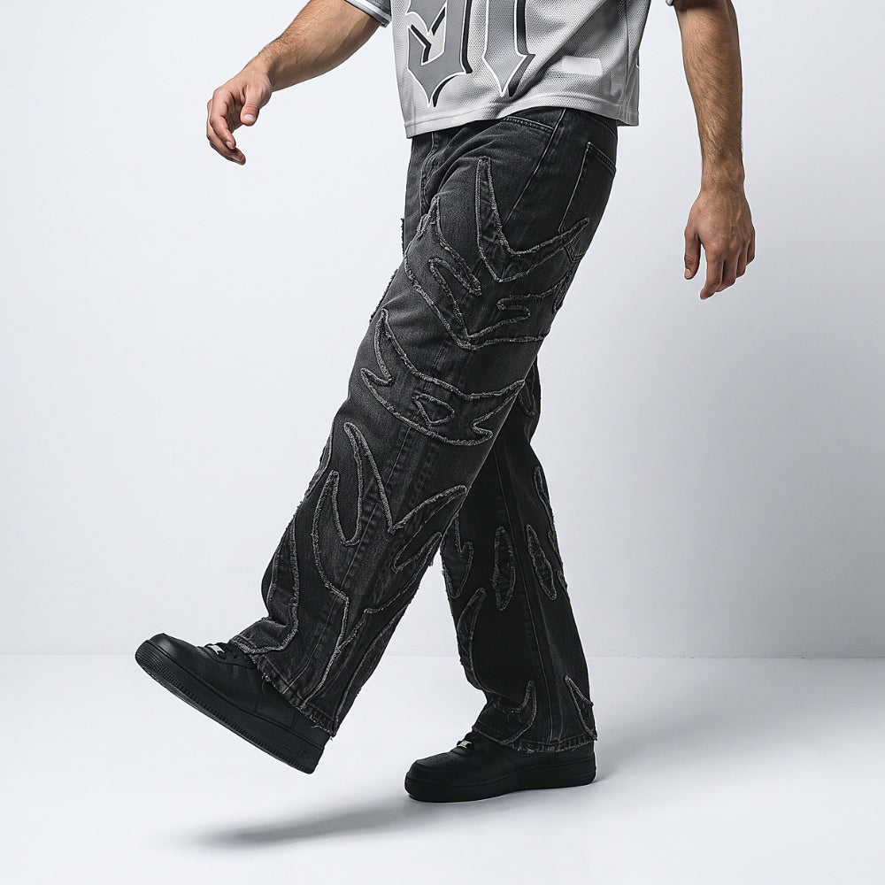 A person wearing black denim jeans with large, abstract white embroidered or printed patterns, paired with a light gray graphic t-shirt and black sneakers, captured in a side-profile walking pose against a light gray studio background.
