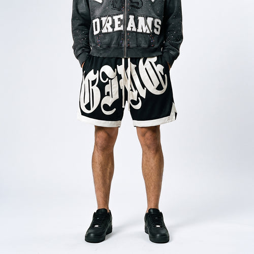Black. A model shown from the waist down wearing black shorts with a large white gothic-style graphic print, a black distressed zip-up jacket, and black sneakers.