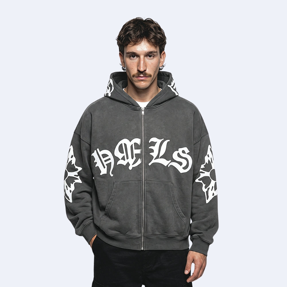 A man wearing a dark gray zip-up hoodie with white gothic lettering and sleeve graphics, photographed against a light gray studio background.