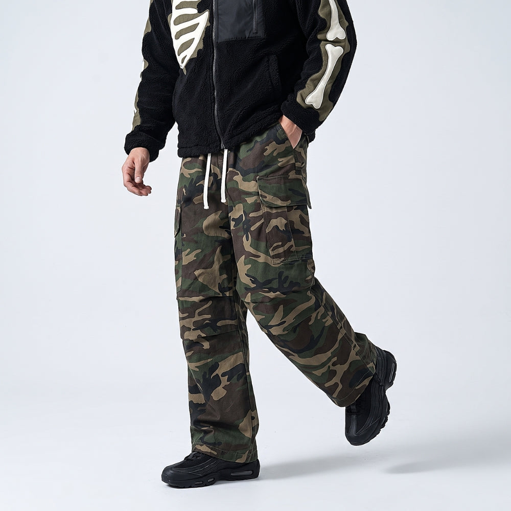 A model wearing a pair of camouflage cargo pants with a black fleece jacket featuring a white skeleton print, captured from the chest down against a light gray studio background.