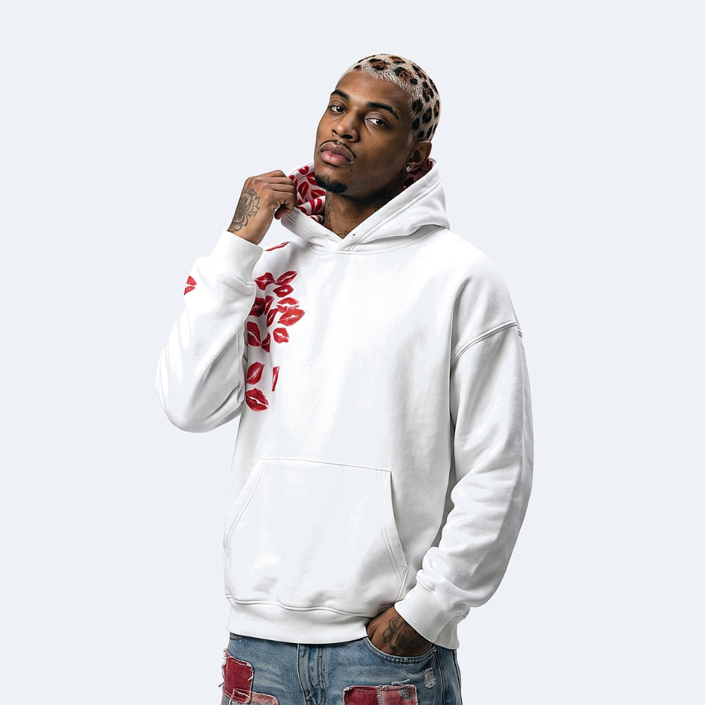 A man wearing a white pullover hoodie with red lip prints on the sleeve and chest, styled with distressed blue jeans and a leopard-print headband, against a light gray background.