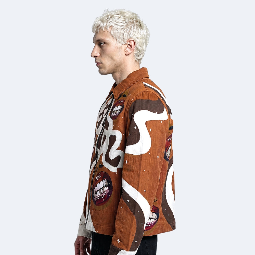 A model with platinum blonde hair shown in profile wearing a brown corduroy button-up shirt with a whimsical graphic print featuring white abstract shapes, cartoonish red mouths, and small white star-like specks, set against a solid light gray background.