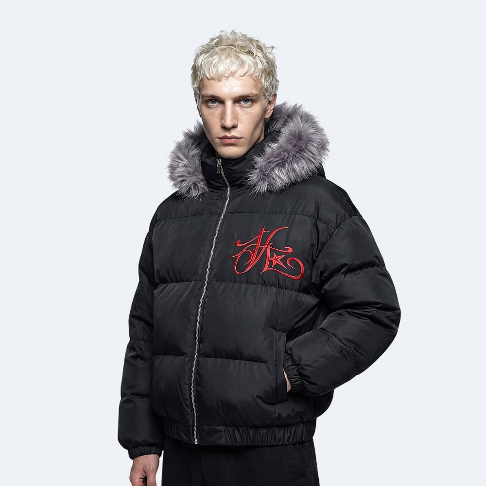 A model with platinum blonde hair wearing a black puffer jacket with a gray faux-fur trimmed hood and a red embroidered logo on the chest, set against a light gray studio background.