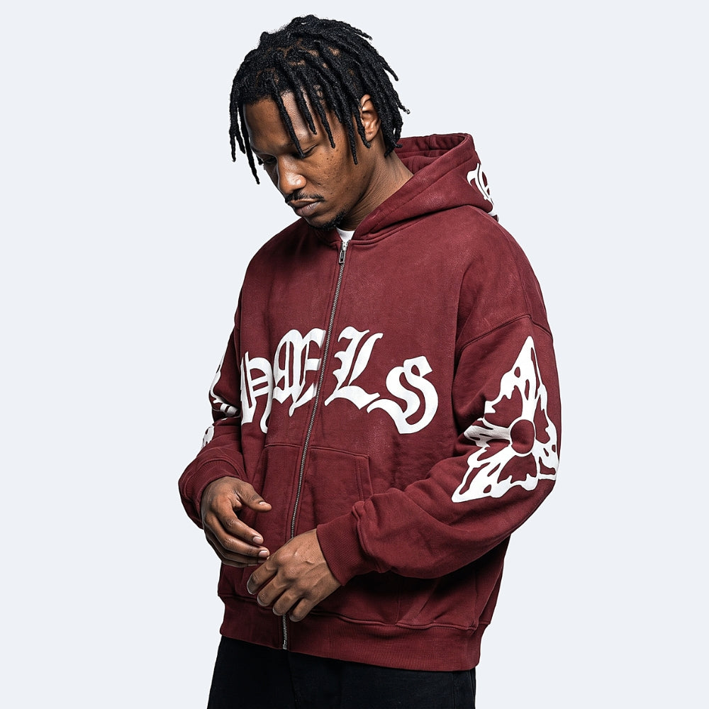 A man wearing a burgundy zip-up hoodie with white Gothic lettering and sleeve graphics, photographed against a light gray studio background.