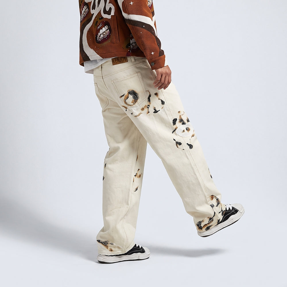 A person wearing cream-colored denim jeans with black and brown cat prints, styled with a brown and white patterned jacket and black sneakers, shown from a side/back angle against a light gray studio background.