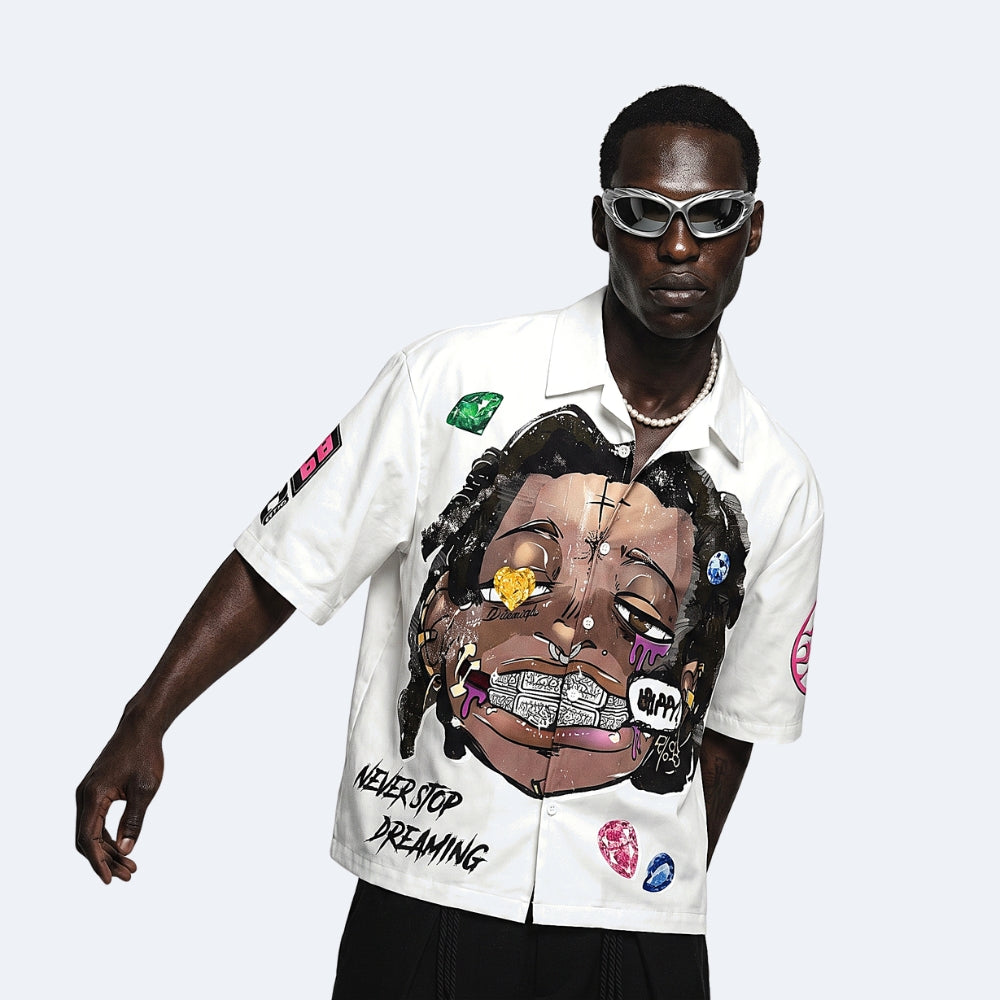 A man modeling a white short-sleeve button-up shirt with a large multicolor graphic print of a face on the front, accompanied by the text 'NEVER STOP DREAMING' and various colorful patch details on the sleeves, set against a light gray background.