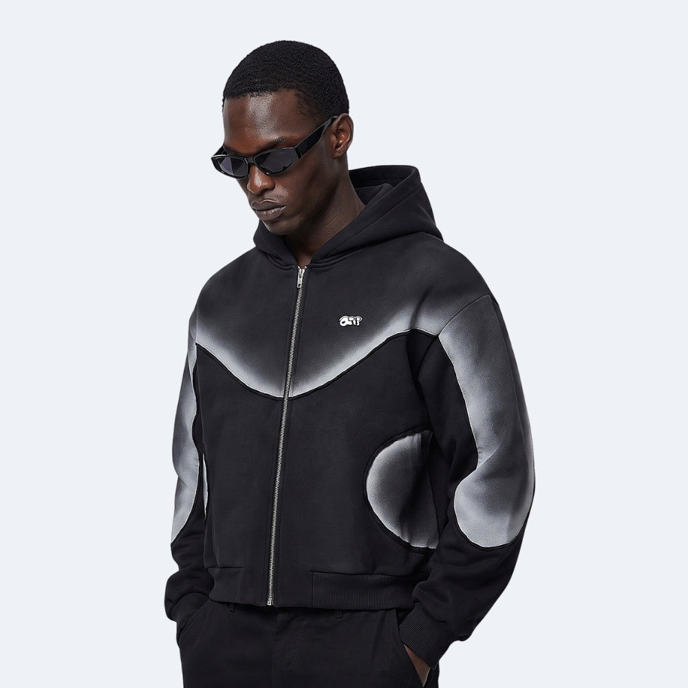A man wearing a black zip-up hoodie with a gray abstract wave pattern on the chest and sleeves, paired with black sunglasses, set against a light gray background.