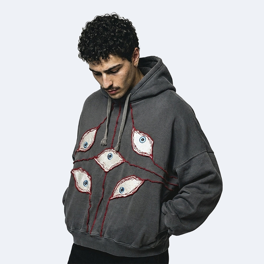 A man wearing a dark gray oversized hoodie with embroidered eyes and red stitched details on the front, photographed against a light gray studio background.