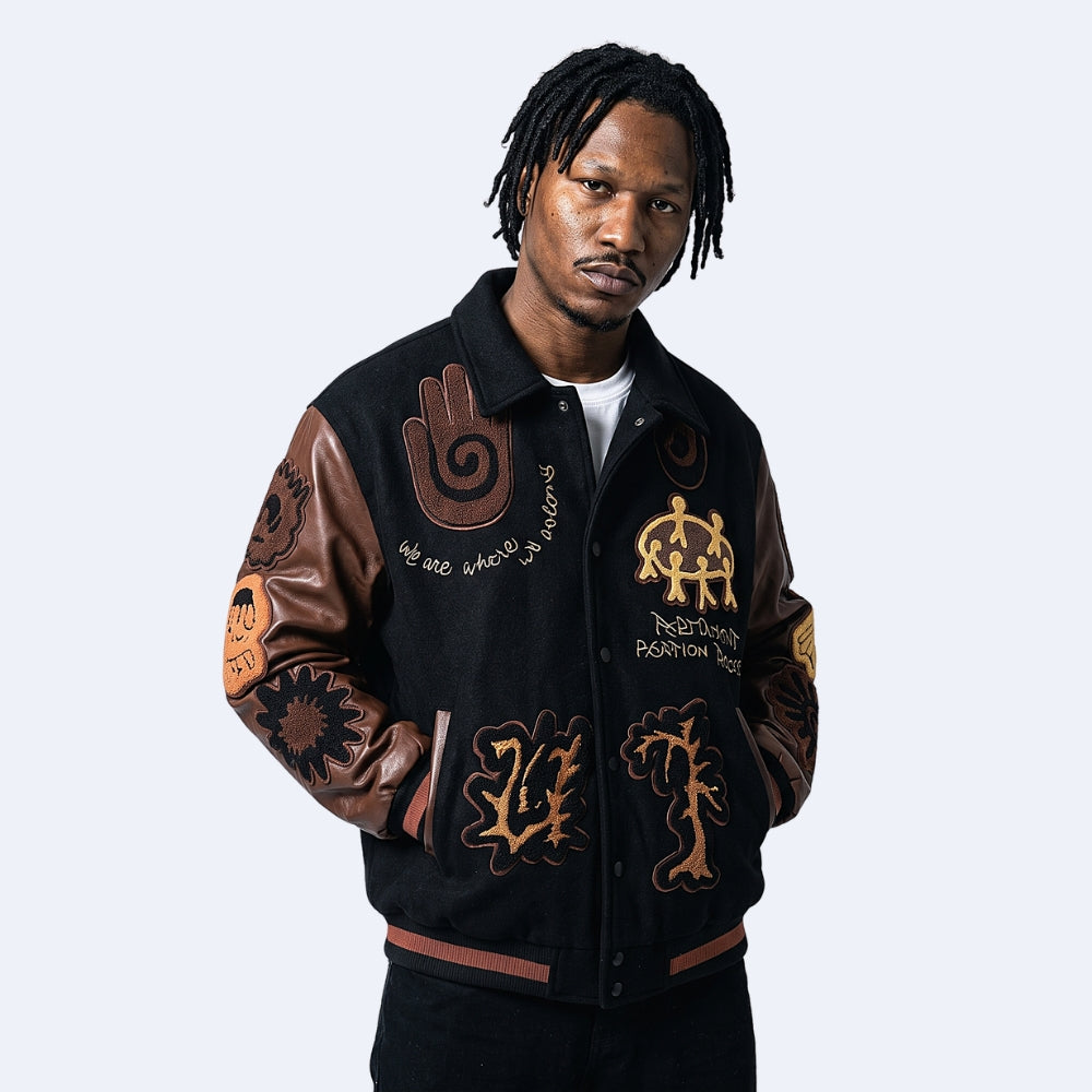 Black. A model wearing a black varsity-style jacket with leather sleeves and various colorful graphic patches.