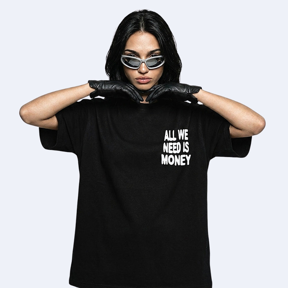 A model wearing a black oversized t-shirt with the text 'ALL WE NEED IS MONEY' printed in white on the chest, styled with black gloves and futuristic silver sunglasses against a light gray background.