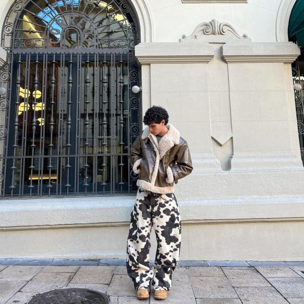 Person wearing a brown shearling jacket and camouflage pants standing in front of a building entrance.
