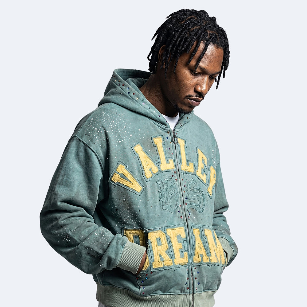 Cyan. A man wearing a distressed sage-green zip-up hoodie with yellow text and studs.