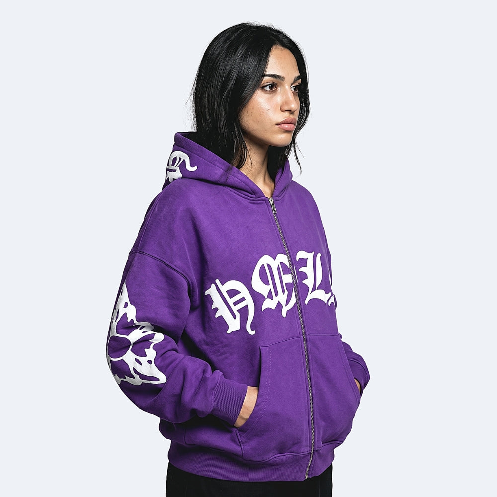 A woman wearing a purple zip-up hoodie with white Gothic lettering and sleeve graphics, photographed against a light gray studio background.