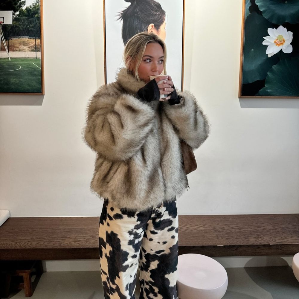 Person wearing a fur coat and patterned pants in an indoor setting with artwork on the wall.