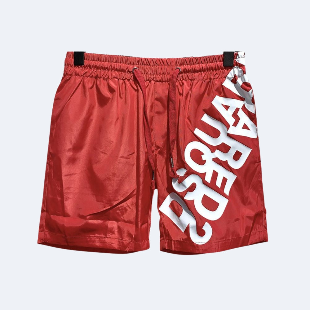 Red. Red athletic shorts with a large white diagonal text print on the left leg, featuring an elastic waistband and black clips at the top.