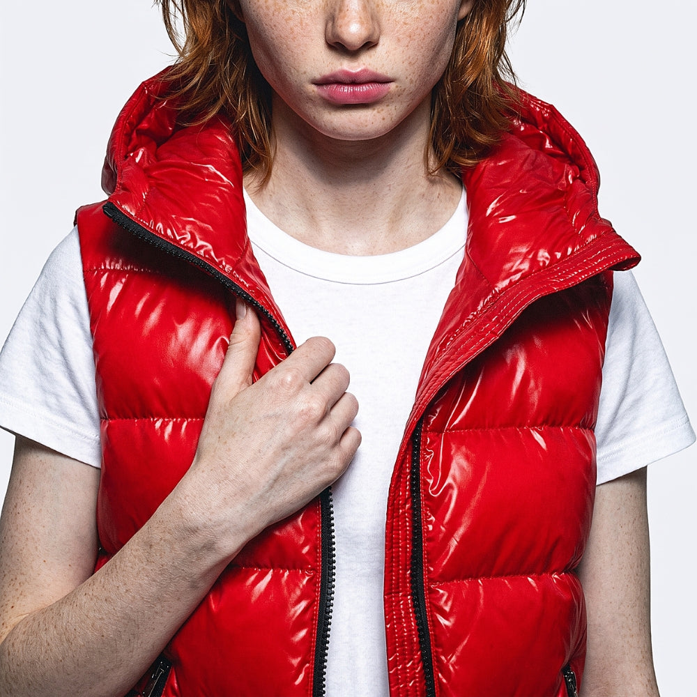 A close-up of a model wearing a shiny, bright red puffer vest over a white t-shirt, with the model's hand adjusting the zipper.